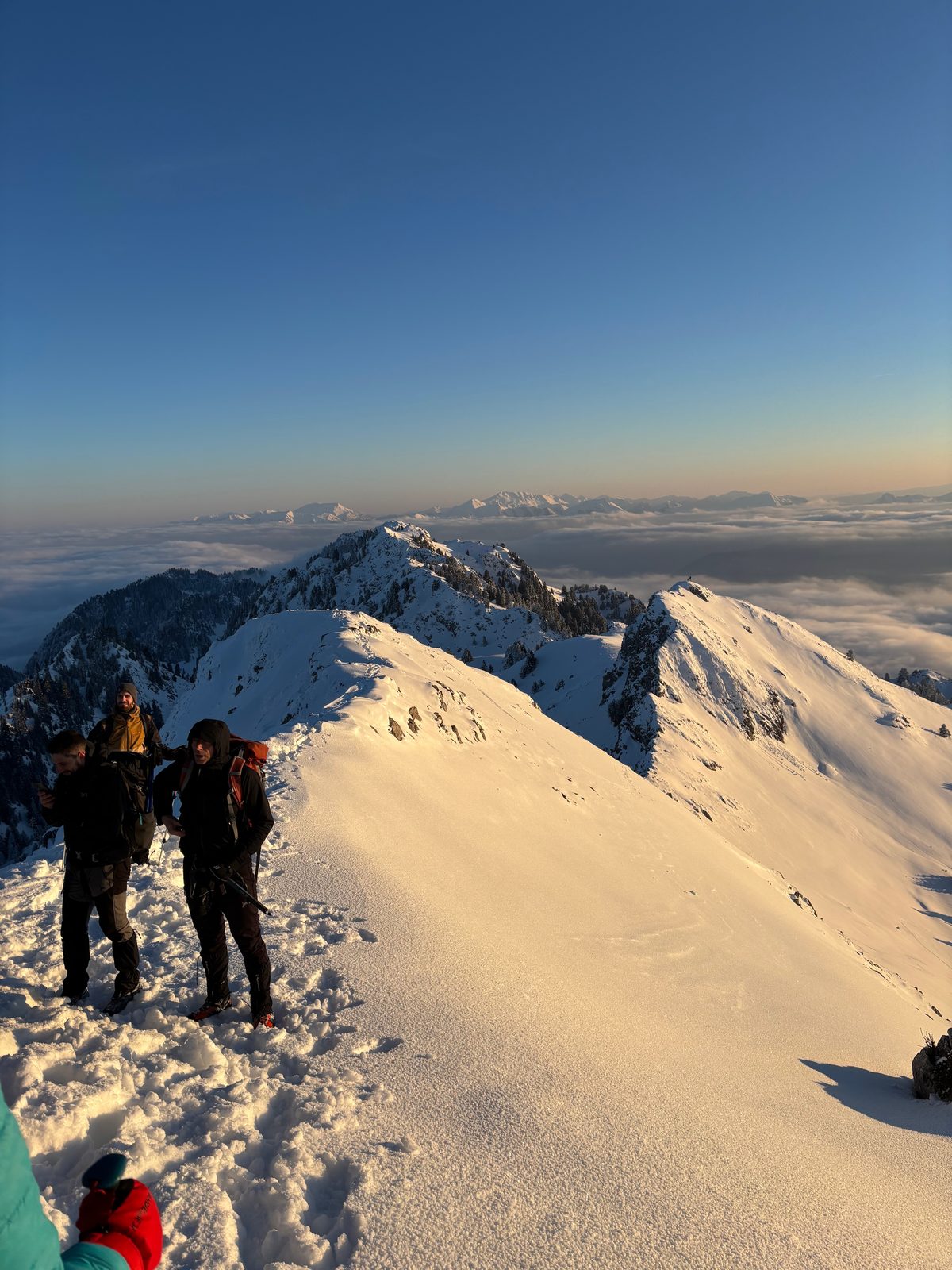 Climbers on a snowy ridge at sunset above a sea of cloud, distant peaks on the horizon