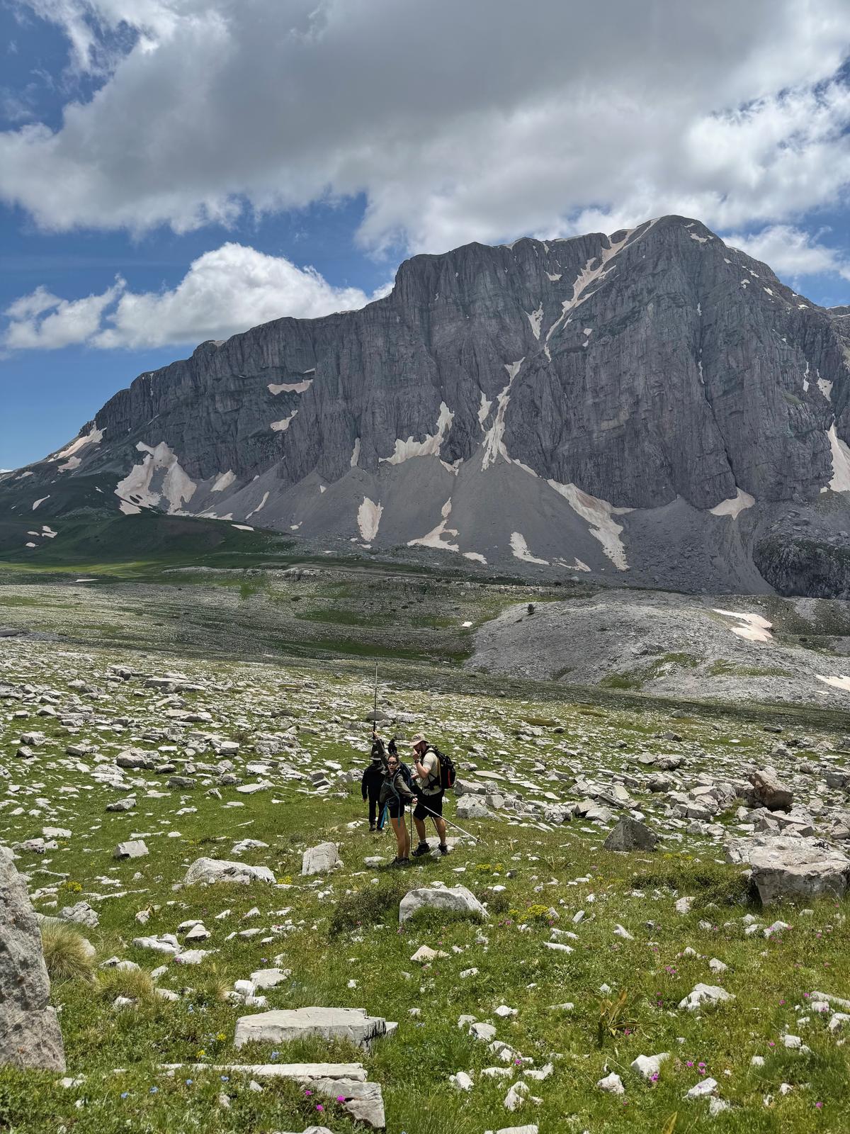 Two hikers crossing a boulder-strewn meadow beneath a large limestone face